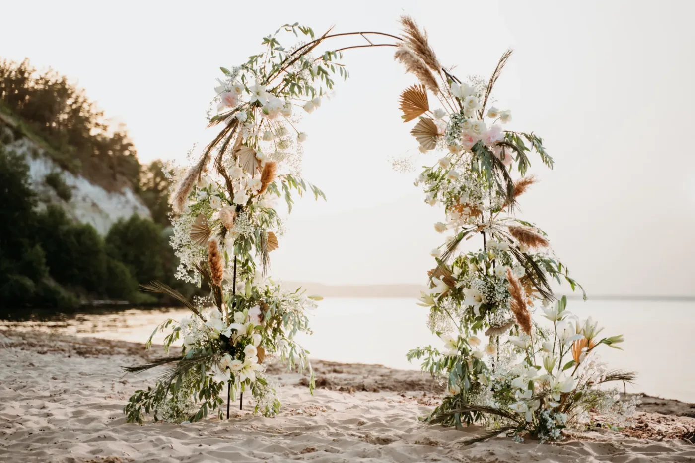 Arcadă florală circulară pe plajă, la apus, decorată cu trandafiri albi și pampas
